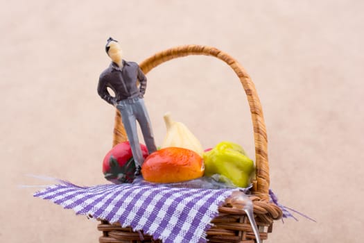 Tiny man figurine on a wicker basket with a plastic fruit on a white and wooden background