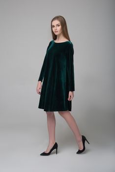 Young beautiful long-haired female model poses in dark green short dress on grey background