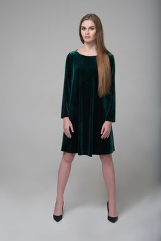 Young beautiful long-haired female model poses in dark green short dress on grey background