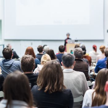 Female speaker giving presentation in lecture hall at university workshop. Audience in conference hall. Rear view of unrecognized participant in audience. Scientific conference event.