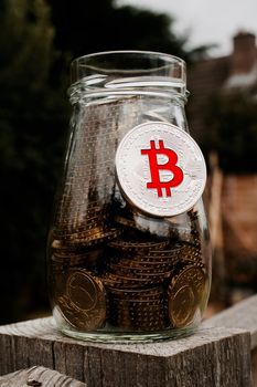 Digital currency physical silver bitcoin coin with red sign on glass jar.