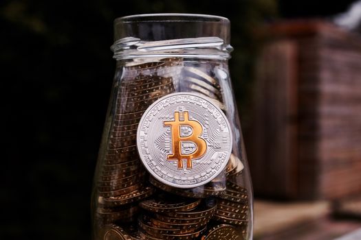 Digital currency physical silver bitcoin coin with yellow sign in glass jar.
