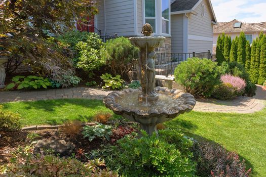 Water Fountain and brick garden path along manicuredhouse frontyard green grass lawn with plants trees and shrubs