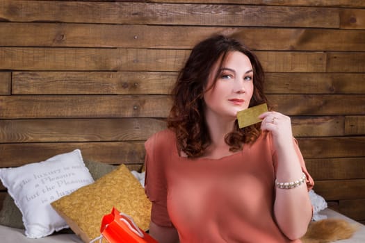 Happy smiling woman after shopping with colourful paper bags and banking credit card sits on bed with wooden wall on background