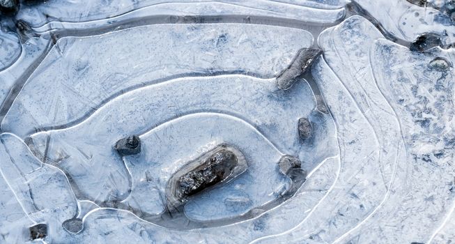 Mud puddle frozen over with white ice crystals, air rings, and rocks visible