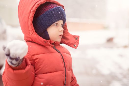 Infant boy while snowing looks towards the emptiness, holding in his hand a snowball, covered with red winter jacket and woolen hat, close-up.