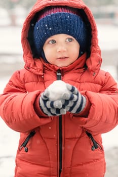 Beautiful infant boy while it is snowing looks towards the void, holding a snowball in his hands, covered with a red winter jacket and a woolen hat, close-up.