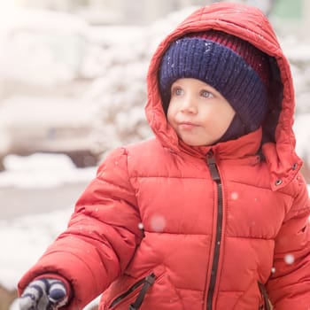 Infant boy while snowing looks towards the emptiness,covered with red winter jacket and woolen hat, close-up.