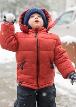 baby boy while it is snowing, looks upwards, holding a snowball in his hand, covered with a winter jacket and a woolen hat, close-up.