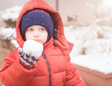 Newborn boy while snowing looks towards the emptiness, holding in his hand a snowball, covered with red winter jacket and woolen hat, close-up.