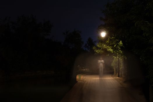 Double exposure night scene of person walking dark street illuminated with streetlights. The receding male silhouettes on the road in the park. Human figure in motion blur going along the city river. Long shutter.