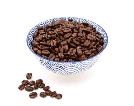 Dark roasted coffee beans in a blue and white ceramic bowl, some spilled beside, on a white background