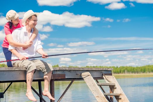 on a wooden pier, a loving couple with a fishing rod is fishing in the lake on a sunny day