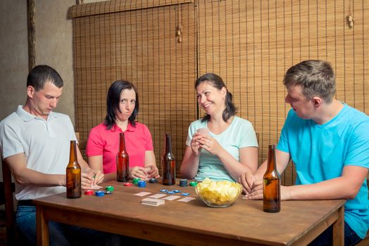 four friends playing cards sitting at a table indoors, drinking beer