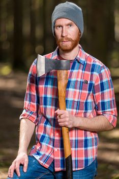 a forester with a red beard posing in a forest with an ax