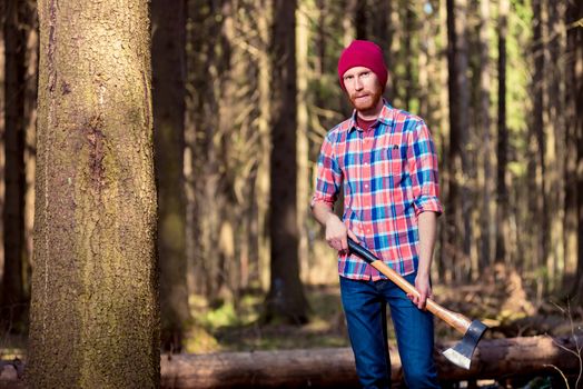 bearded logger in a checkered shirt with an ax standing near a tree in the forest