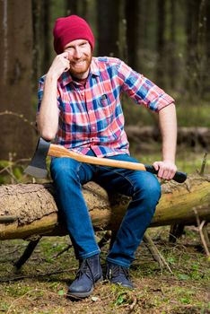 happy bearded lumberjack with an ax is sitting on a log in the forest