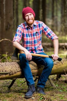 a smiling bearded lumberjack in a hat and shirt with an ax sits on a log in the woods
