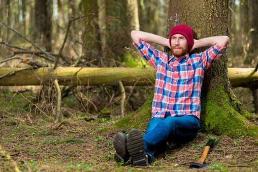 dreamy and tired bearded forester with an ax on the relaxation in the forest