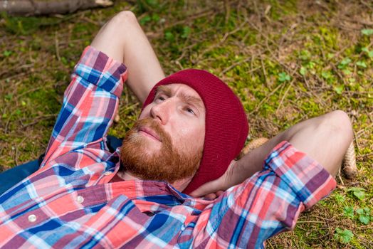 portrait of a hipster with a beard in a red hat, resting in the forest on the ground