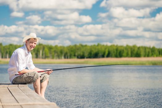 happy fisherman in a hat on a wooden pier on a beautiful lake with a fishing rod