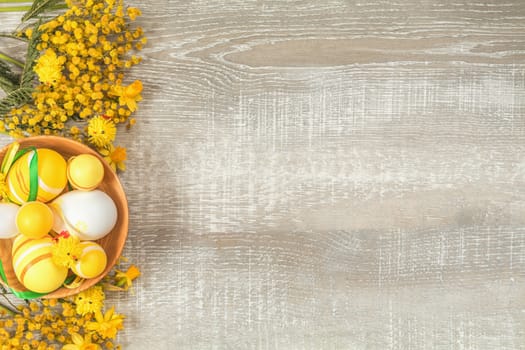 Composition with painted eggs in a wooden bowl. Easter accessories, mimosa and yellow daffodils on a light wooden surface. Yellow orange easter concept, top view, copy space,  in yellow tones on a light wooden surface