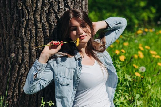 Young woman smelling dandelion flower sitting on a lawn under a tree in park