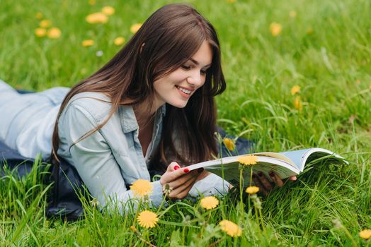 Beautiful young woman reading a book on grass with dandelion flowers in park