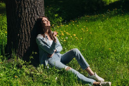 Young woman smelling dandelion flower sitting on a lawn under a tree in park