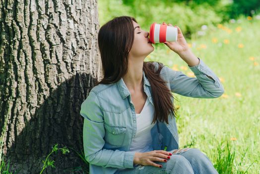 Beautiful girl enjoying a cup of coffee sitting under tree in park