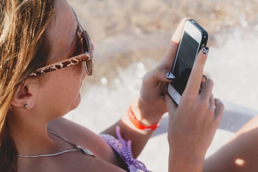 A young tanned woman looks into a smartphone through sunglasses on a lounger. The concept of a lifestyle is always on the Internet.