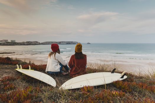 Two best friends sitting near the coastline with her surfboards while looking to the ocean