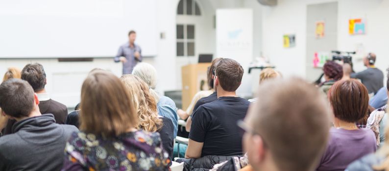 Male speaker giving presentation in lecture hall at university workshop. Audience in conference hall. Rear view of unrecognized participant in audience. Scientific conference event.