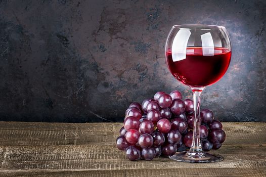 Wineglass with red wine and red grapes on wooden table. Dark background.