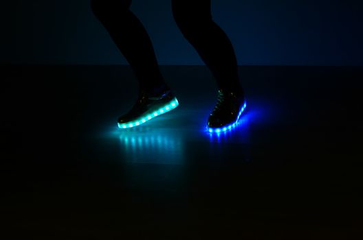 Fashionable sneakers with neon LED lighting on the legs of a girl with blue and azure colors in the dark .Different colors of neon lights soles sneakers are present.