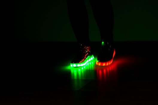 Fashionable sneakers with neon LED lighting on the legs of a girl with red and green colors in the dark .Different colors of neon lights soles sneakers are present.