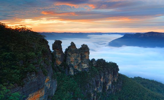 Beautiful sunrise over the famous landmark, the Three Sisters with a dense fog in the Jamison Valley floating like a river.  Location Echo Point, Katoomba, Blue Mountains, Australia