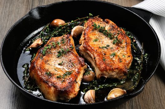Juicy, browned pork steak on a bone in oil with garlic and herbs in a frying pan. Close-up.