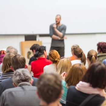 Male speaker giving presentation in lecture hall at university workshop. Audience in conference hall. Rear view of unrecognized participant in audience. Scientific conference event.