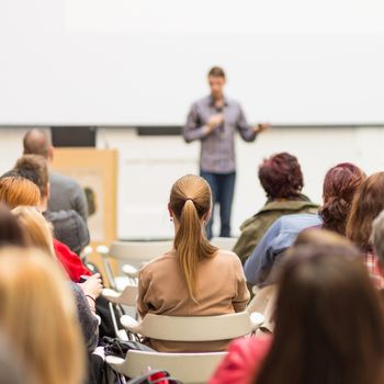 Male speaker giving presentation in lecture hall at university workshop. Audience in conference hall. Rear view of unrecognized participant in audience. Scientific conference event.