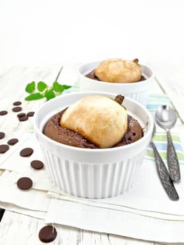 Two white bowl dessert with chocolate and pear, spoon, mint on a kitchen towel on the background light wooden boards