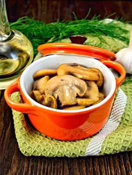 Champignons fried in a red ceramic pot on a napkin, garlic, dill and vegetable oil on the background of wooden boards