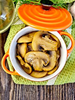 Champignons fried in a red ceramic pot on a napkin, garlic, dill and vegetable oil on the background of wooden boards on top