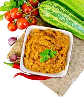 Caviar squash in a clay bowl on a napkin of burlap, hot peppers, tomatoes, zucchini, parsley and garlic isolated on white background on top