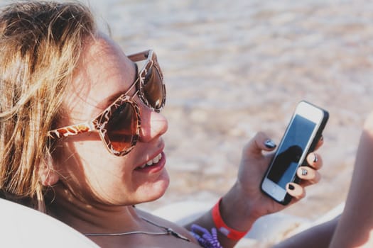 A young tanned woman looks and laughs into a smartphone through sunglasses on a lounger. The concept of a lifestyle is always on the Internet.