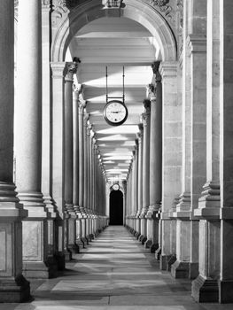 Long colonnafe corridor with columns and clock hanging from ceiling. Cloister perspective. . Black and white image.