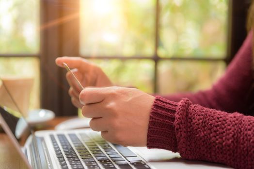 Pretty Young womans hands holding a credit card and using laptop computer for online shopping. Online payment. Female working on laptop in a cafe. Note book, pen and credit card put on table.