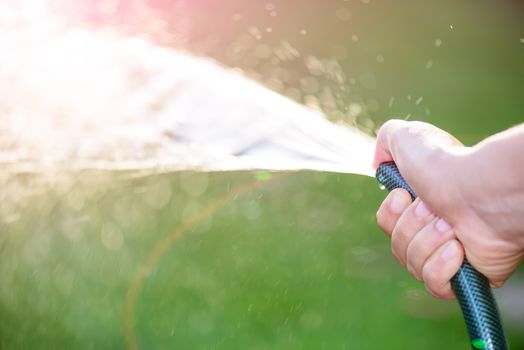 Woman hand holding rubber water hose and using finger close end of rubber water hose to make  water spray with sunlight and green grass field in background. World Water Day conpect.