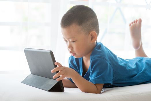 Young child addicted to technology gadget. Asian boy playing with digital tablet pc at home.