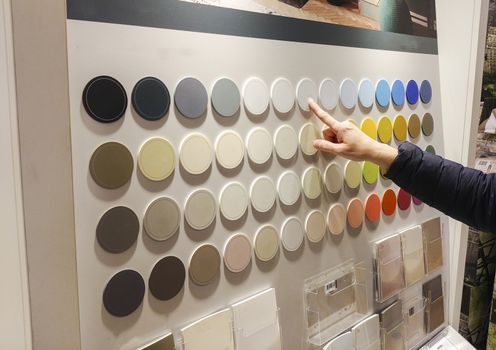a woman in a blue coat pointing with her hand to a certain color of wall paint in a shop with do it yoursaelf products